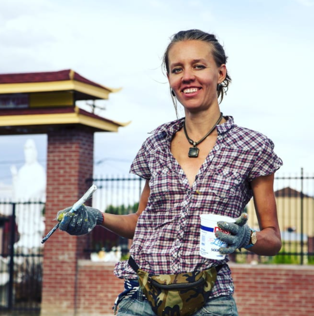 Artist Yulia Avgustinovich working on a mural, holding a paintbrush and paint, smiling at the camera.