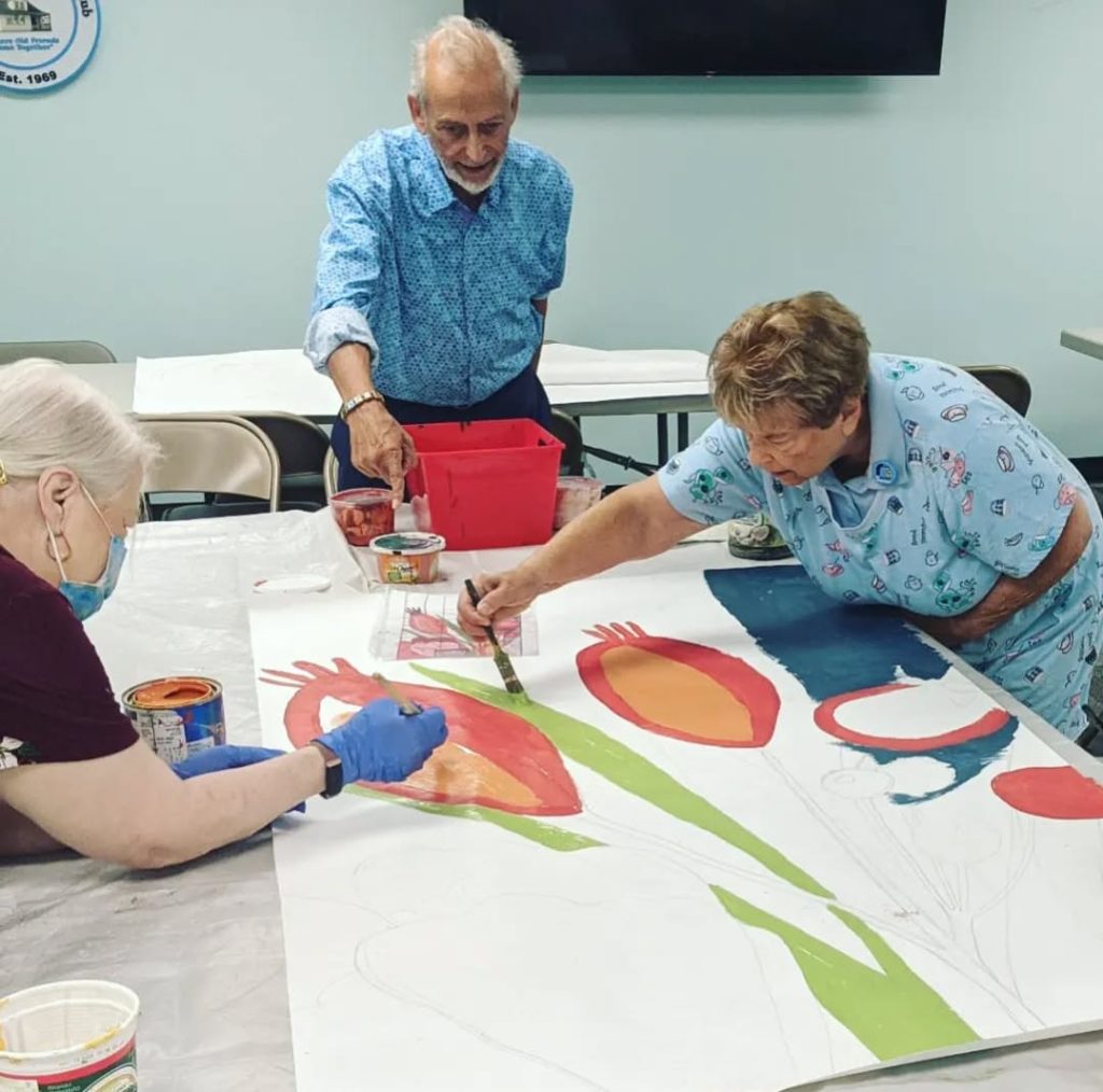 Retired volunteers painting a floral mural fragment on Polytab fabric inside a senior living facility for a community mural in Runnemede, New Jersey, by Yulia Avgustinovich.