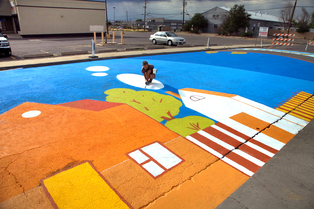 about-professional-muralist-Yulia_Avgustinovich_Aurora_Street_mural_Stanley_Market_Place_Colorado Artist Yulia Avgustinovich working on a huge ground mural, holding a paintbrush and paint in Aurora CO.